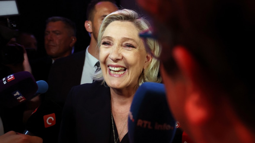A middle-aged blonde woman in a suit laughs while speaking to reporters.