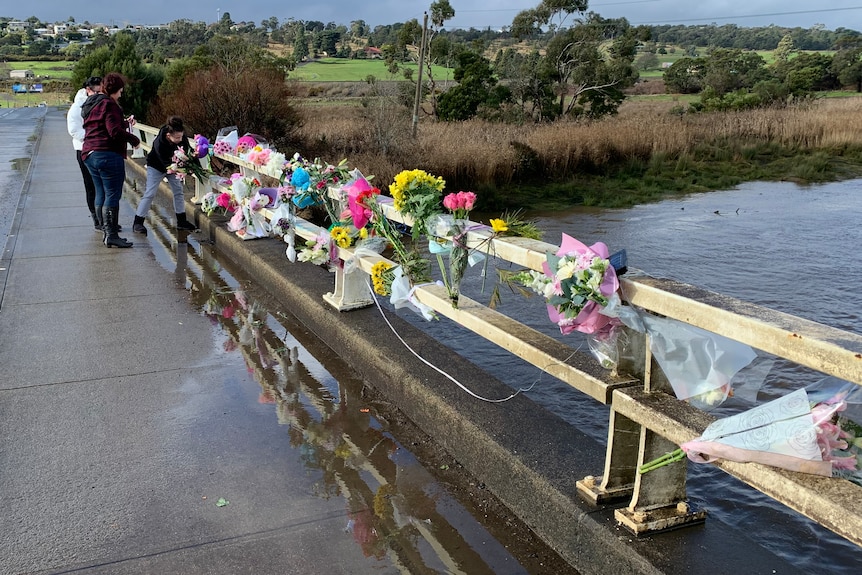 Three people attach a floral tribute among others on a bridge railing.