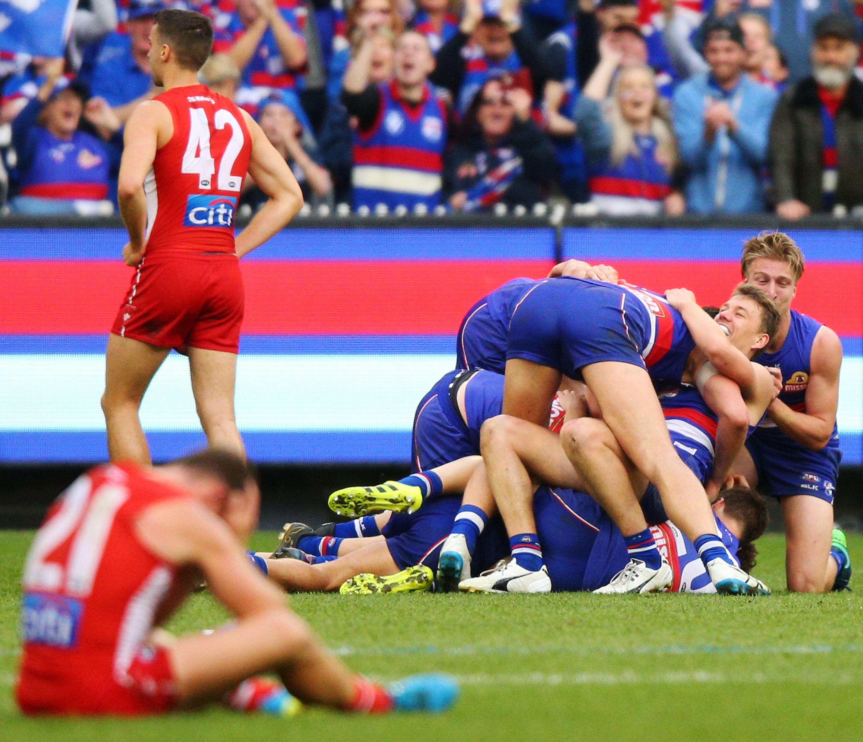AFL Western Bulldogs players pile on top of each other in celebration as dejected Sydney players stand or sit on the ground. 