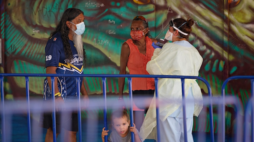 An Aboriginal man and woman in face masks speak with a health worker in a mask and face shield.