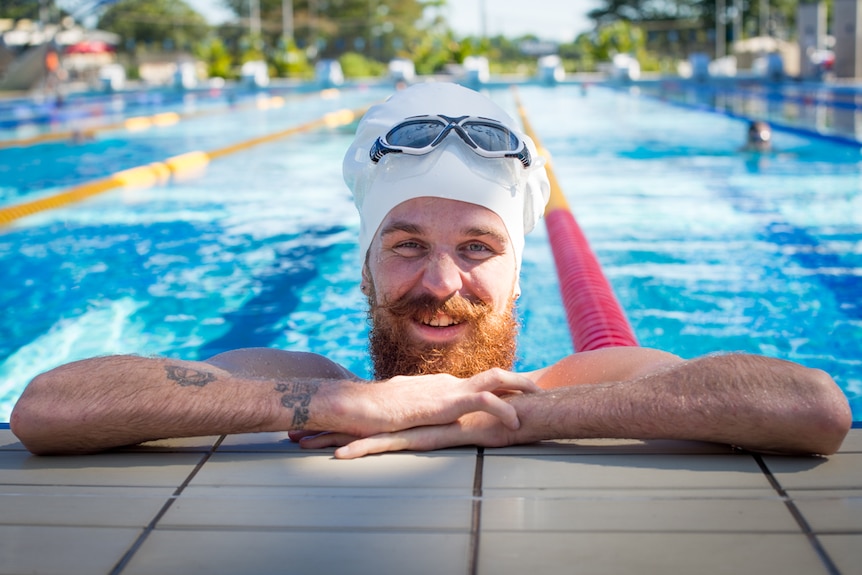 A bearded man in an enlarged swimming cap rests against the side of a pool with his goggles on his head.