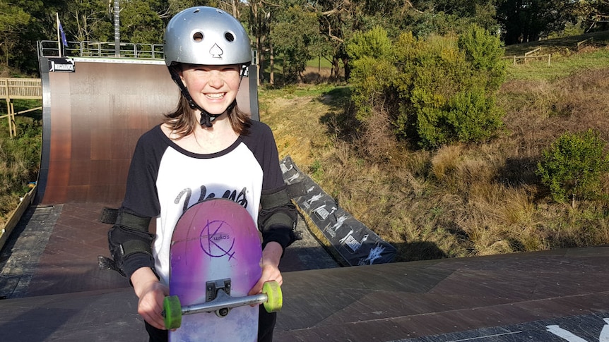 A girl wearing a helmet and holding a skateboard, standing at the top of a giant skate ramp.