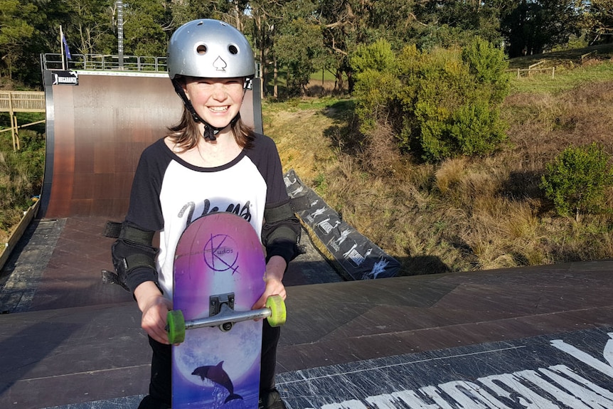 A girl wearing a helmet and holding a skateboard, standing at the top of a giant skate ramp.