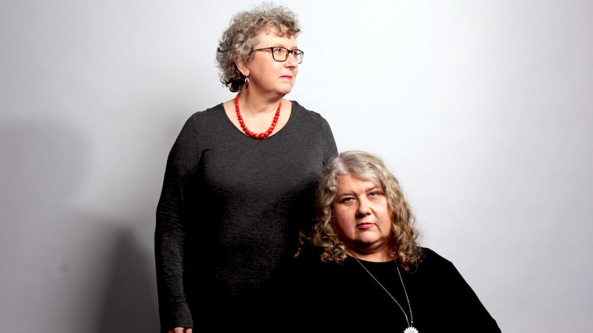 The two woman, Rozanna standing and Kate sitting, pose in front of a grey backdrop, both wearing grey.