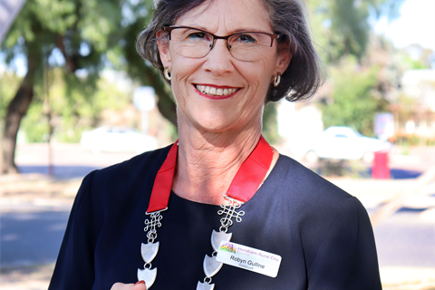 A woman holds a red necklace with a metal chain. She is smiling and wearing glasses. It is sunny.