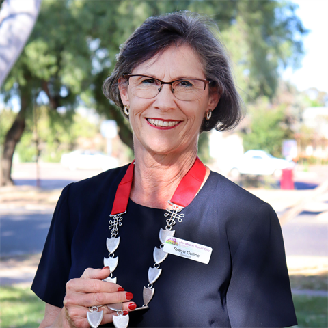 A woman holds a red necklace with a metal chain. She is smiling and wearing glasses. It is sunny.