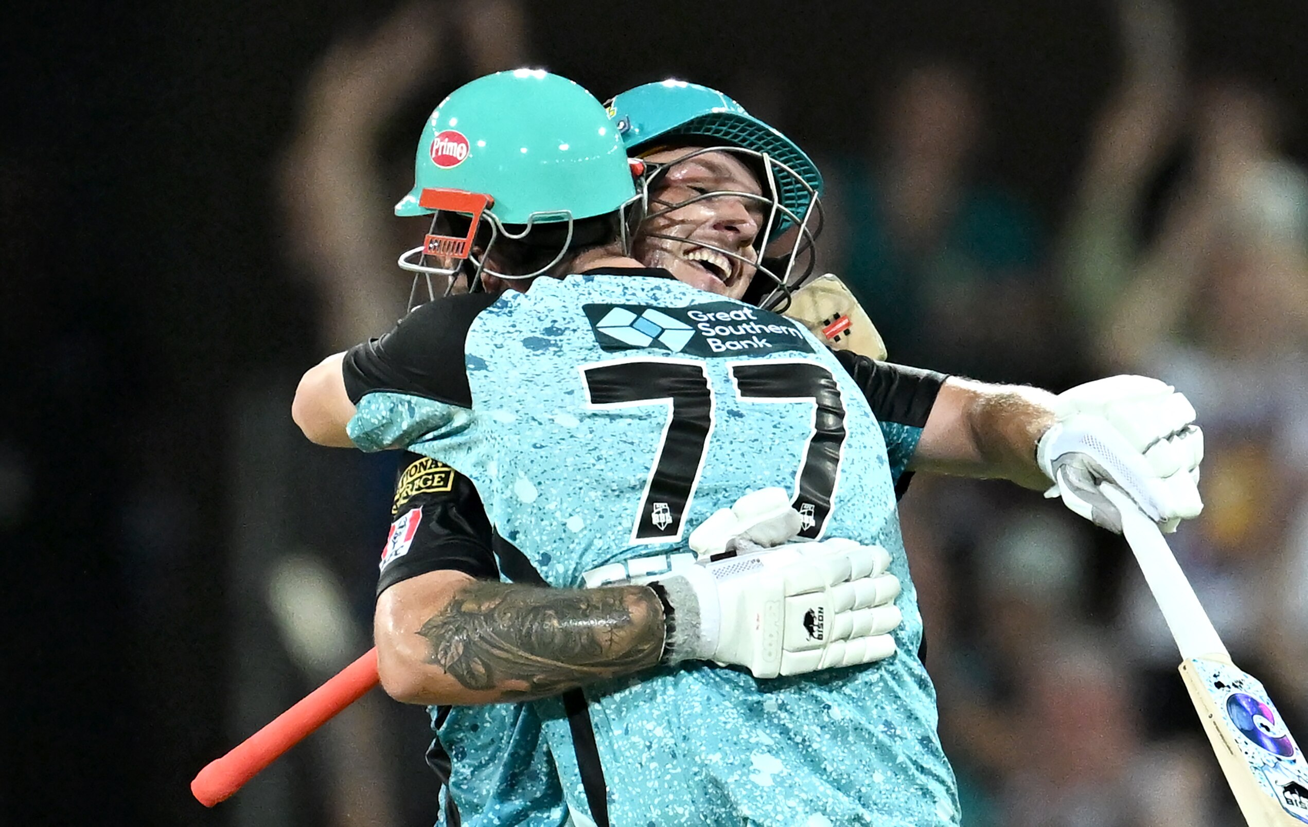 Two male cricketers, batting in the middle, hug each other in celebration.