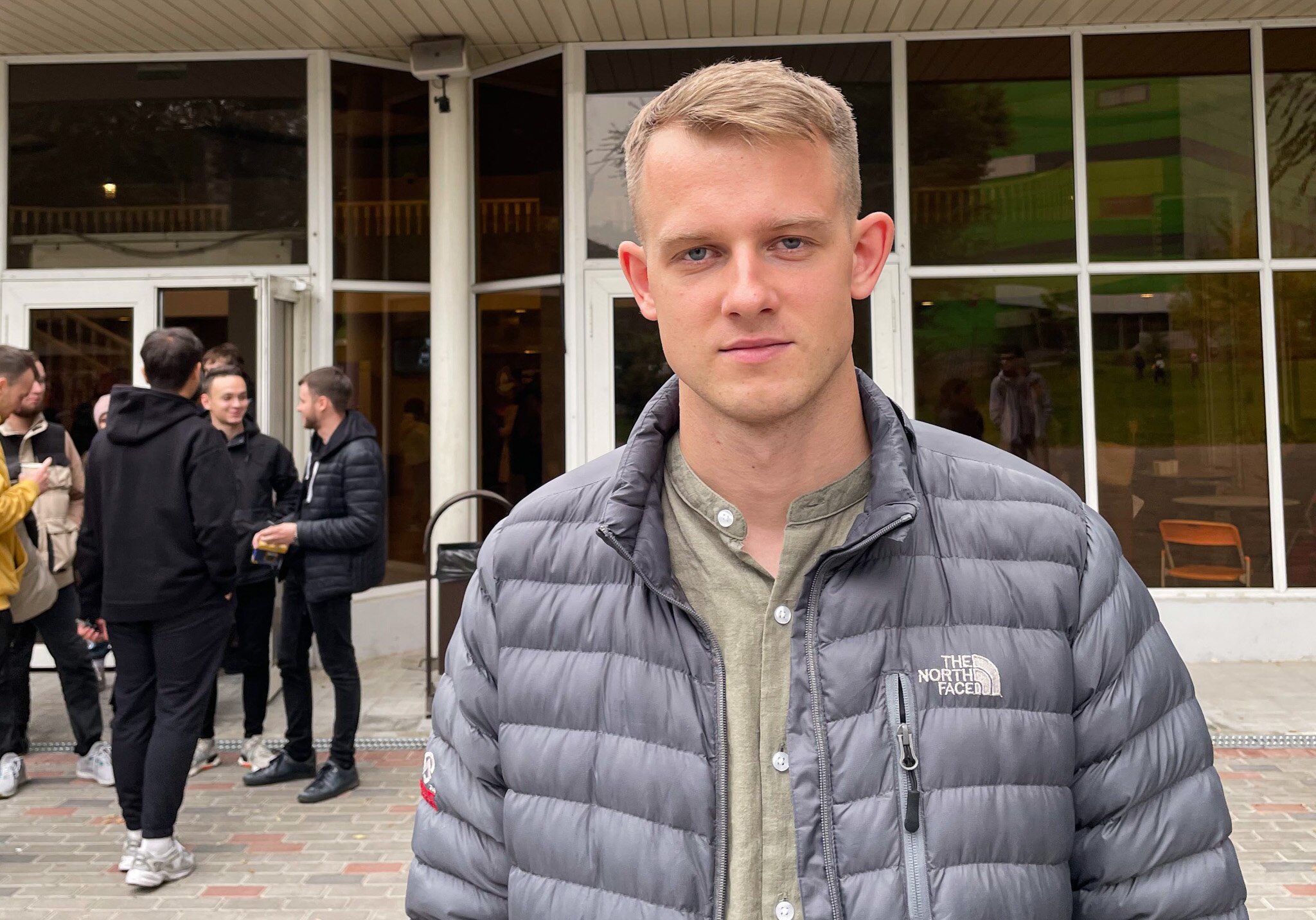 A man with blonde hair and wearing a black puffer jacket stands outside a church with people gathered outside.