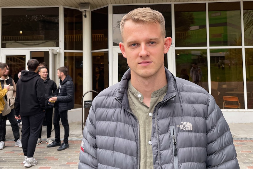 A man with blonde hair and wearing a black puffer jacket stands outside a church with people gathered outside.