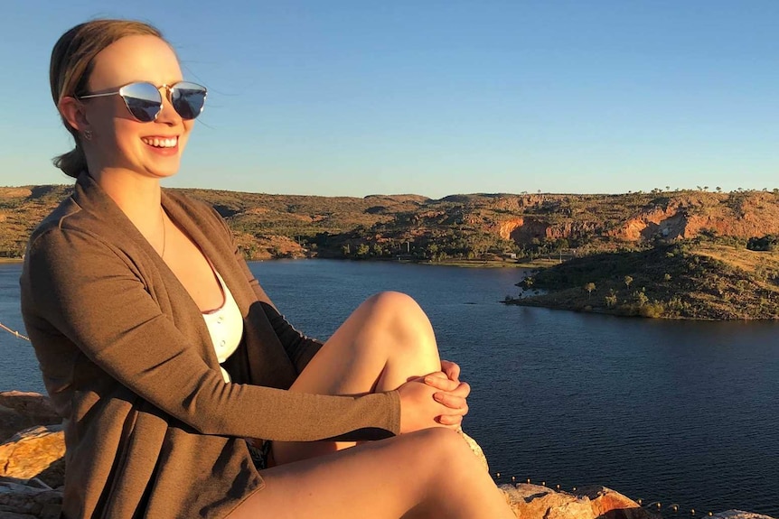 A young woman with blonde hai rand sunglasses sits atop a rock, looking out over a lack in the outback.