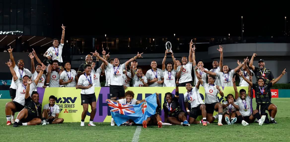Group of Fijian women wearing white rugby jerseys celebrate winning by raising a trophy, holding Fiji flag with their hands up