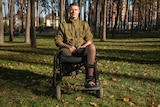 A man in a green army jacket sits in a wheelchair in a clearing in a wood 