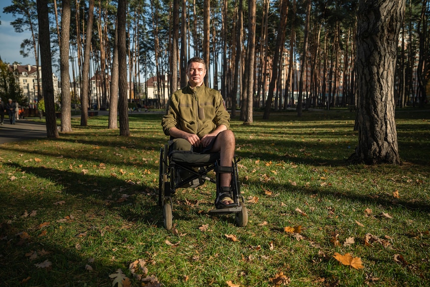A man in a green army jacket sits in a wheelchair in a clearing in a wood 