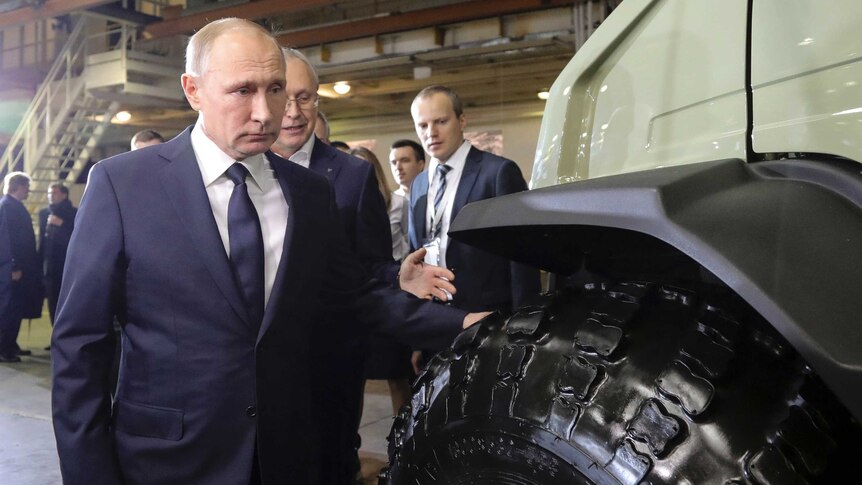 Russian President Vladimir Putin inspects the wheel of a large vehicle.