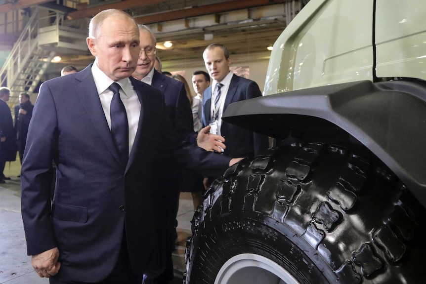 Russian President Vladimir Putin inspects the wheel of a large vehicle.