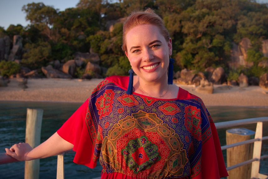 A woman smiles while holding the rail of a boat with the beach in the background