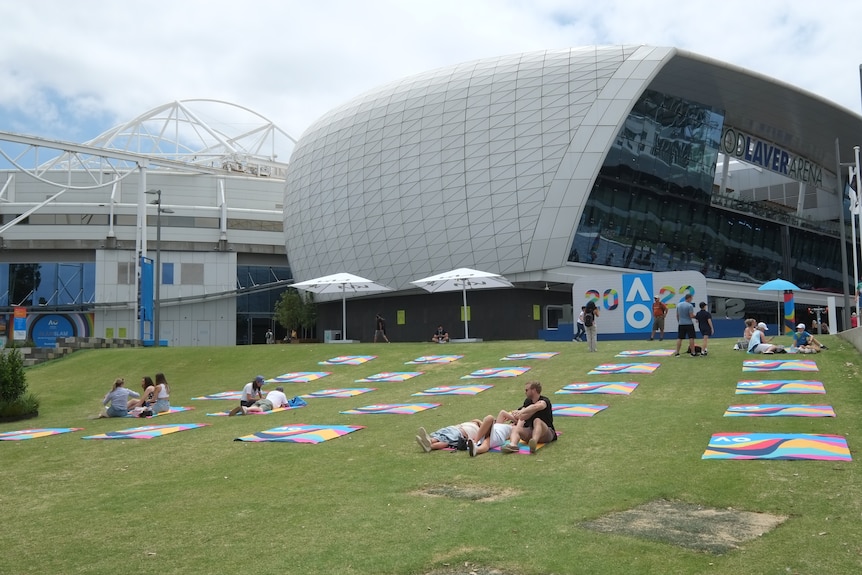 Grass in front of Rod Laver Arena with colourful mats laid out about a metre apart, some people lying on them.