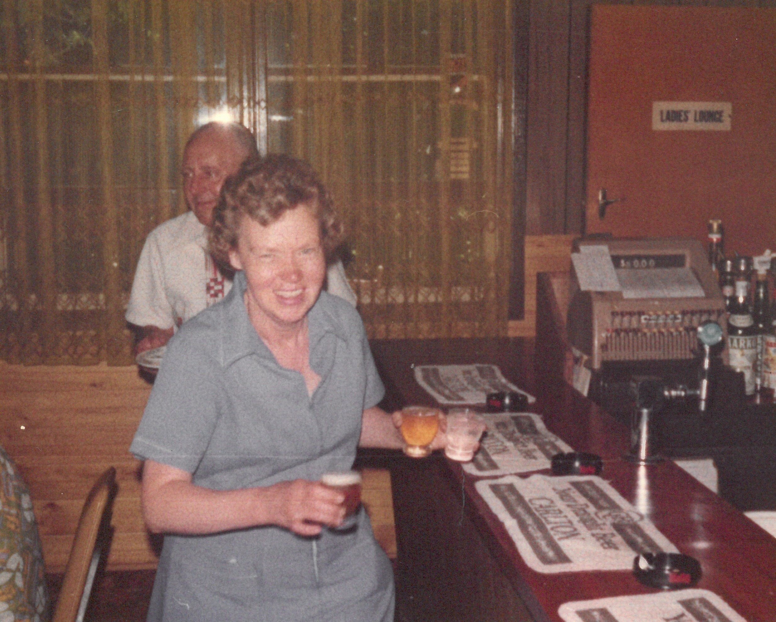 A 1970s photo of an older woman at a bar, holding three drinks, with a man in the background