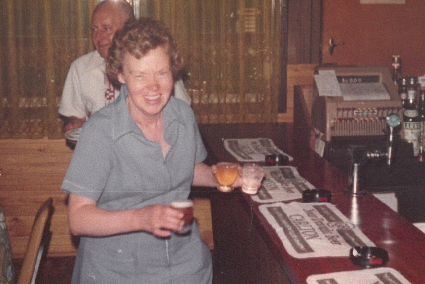 A 1970s photo of an older woman at a bar, holding three drinks, with a man in the background