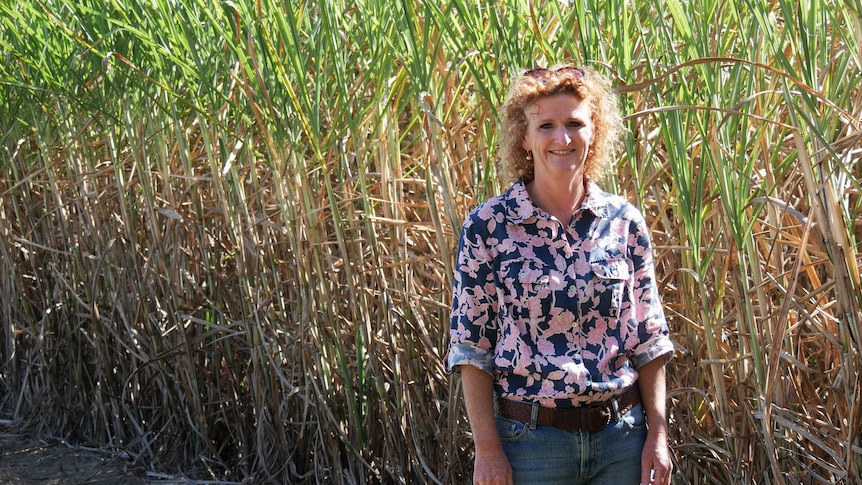 Judy Plath, cane grower with red curly hair, purple and pink shirt stands in front of cane field