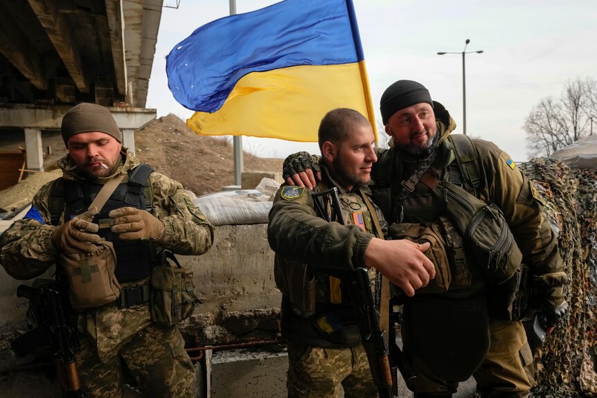 Three Ukrainian soldiers stand together in front of a Ukrainian flag