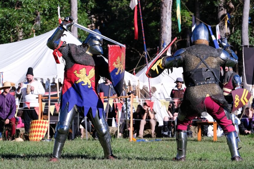 Two men wearing full body armour fight with sword and shield.