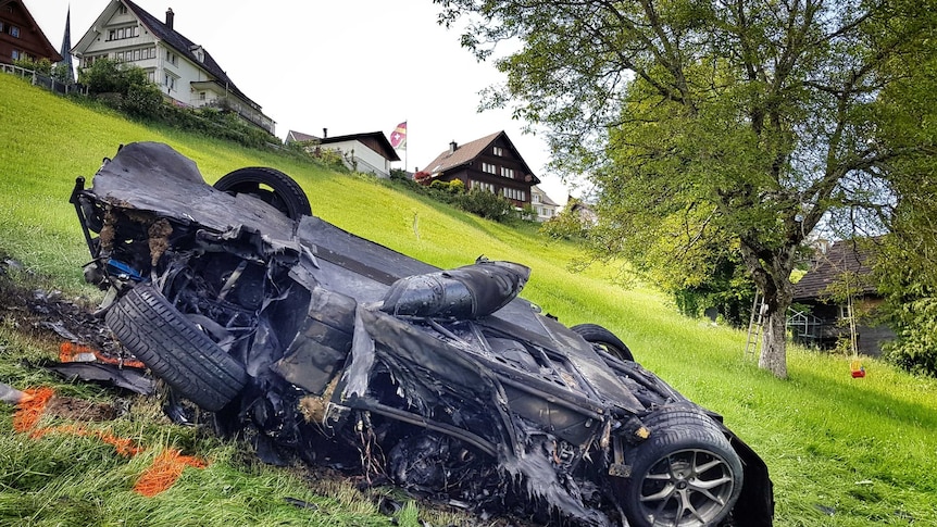 A blackened, twisted car on a green hillside in Switzerland, with houses in the background.