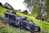 A blackened, twisted car on a green hillside in Switzerland, with houses in the background.