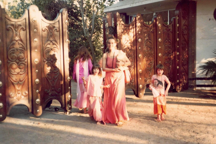 A woman and two girls dressed in orange stand at the gates of a commune.
