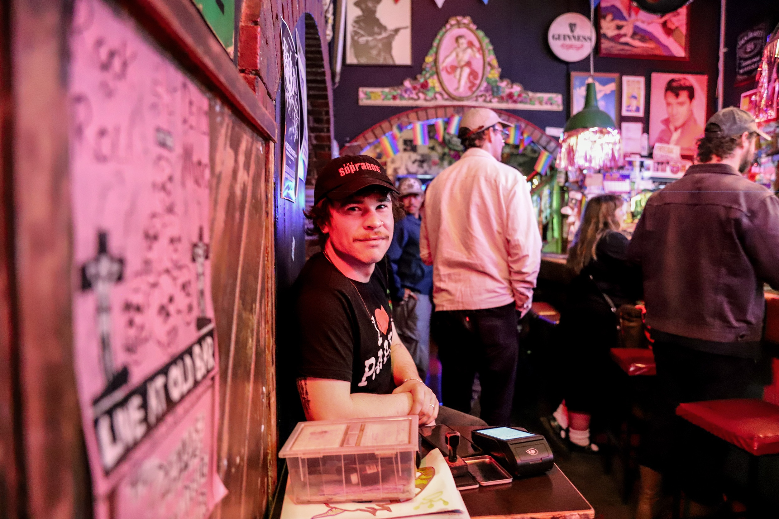 Homem de boné sorri sentado atrás de uma mesa em um bar lotado