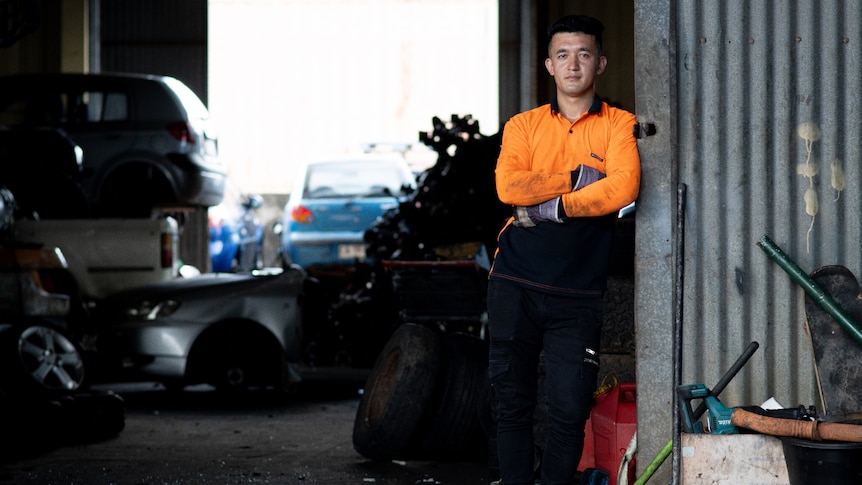 A wide shot of Abbas Haidar, wearing high vis and leaning against the doorway of a mechnical warehouse.