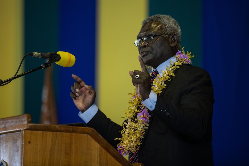 A man wearing a suit and a necklace of flowers speaks behind a podium.