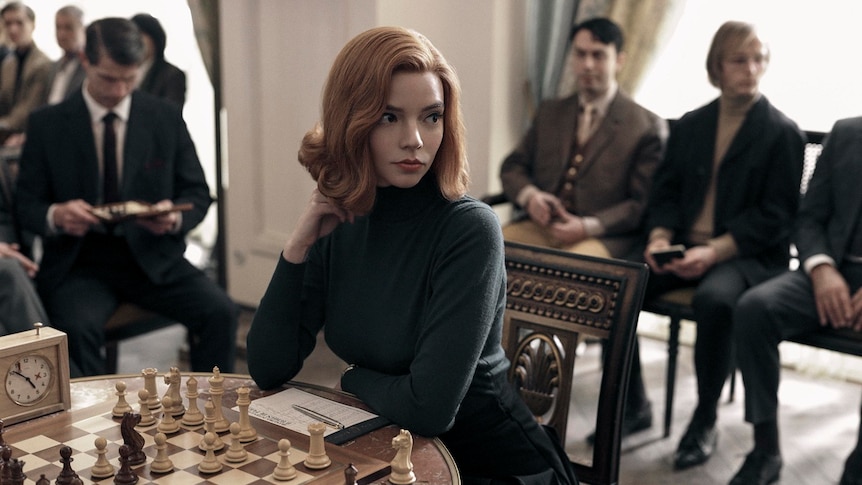 A woman with orange hair wearing a black high neck sweater sitting at a table with chess board and chess pieces