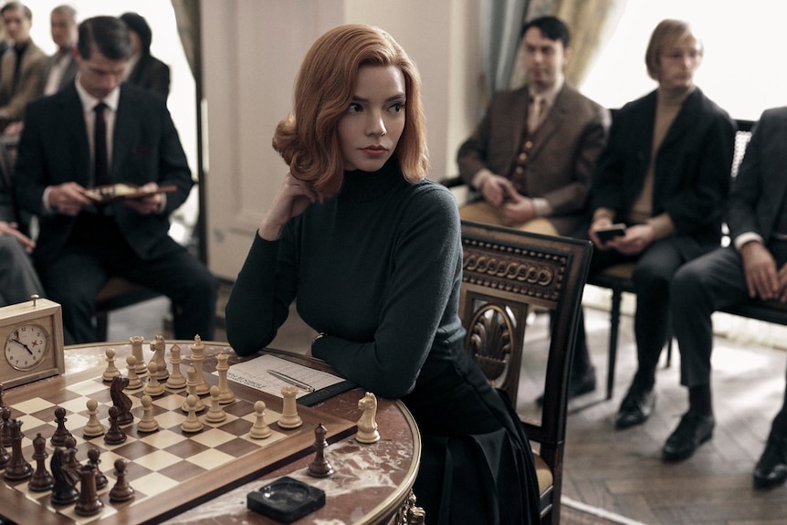 A woman with orange hair wearing a black high neck sweater sitting at a table with chess board and chess pieces