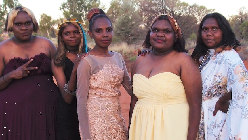 Five Indigenous girls wearing formal dresses pose in a desert landscape