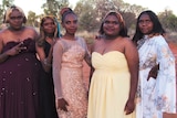 Five Indigenous girls wearing formal dresses pose in a desert landscape