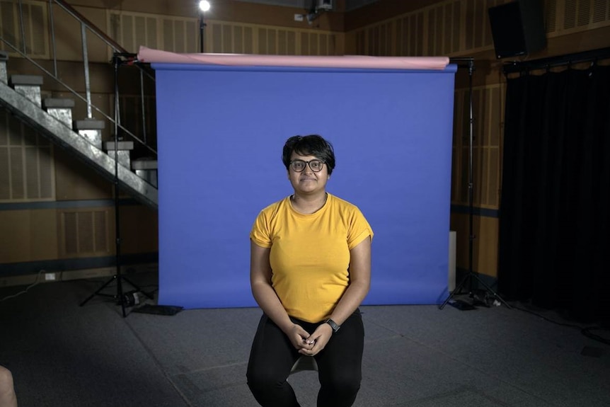 A woman wearing glasses and a yellow top in front of a purple screen in a studio.