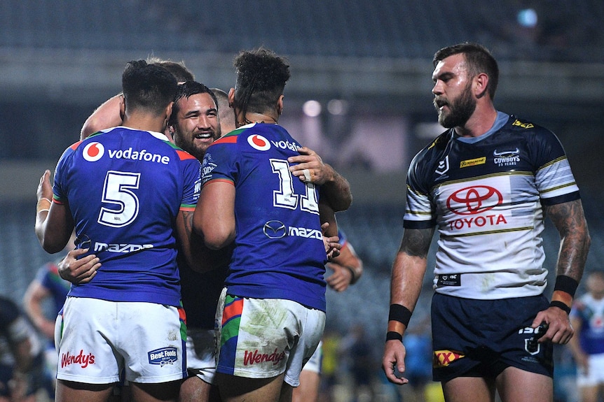 An NRL player smiles as he is surrounded by teammates after scoring a try, as an opponent looks on.