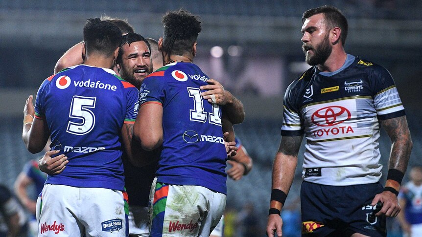 An NRL player smiles as he is surrounded by teammates after scoring a try, as an opponent looks on.