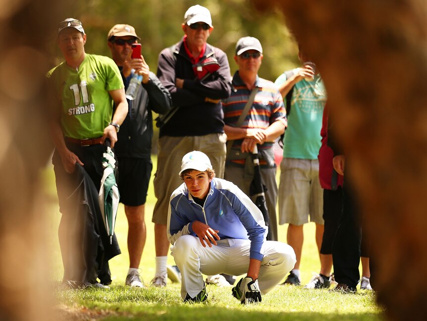 A teenage golfer crouches down to stare through the trees to prepare a shot at the Australian Open with the crowd behind him.
