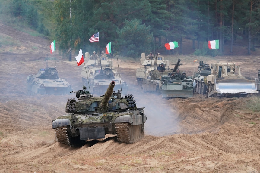 Military vehicles and tanks with flags from from various NATO ocuntries drive across rough dirt roads.