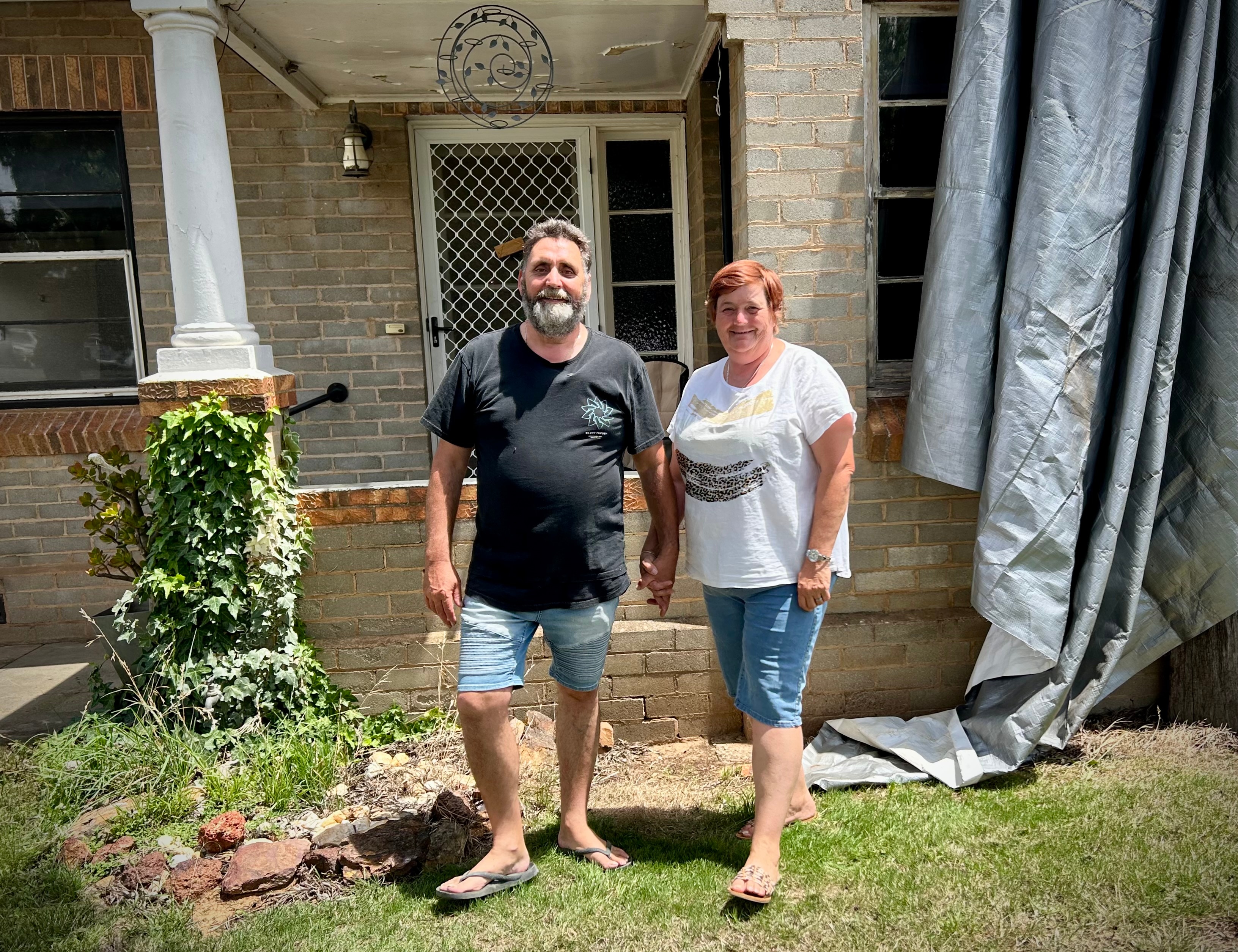 A man and a woman stand in front of a house, with tarp covering the front window.