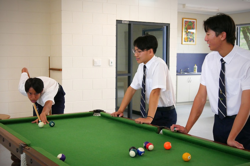 Three young boys in formal school uniform stand around a pool table.