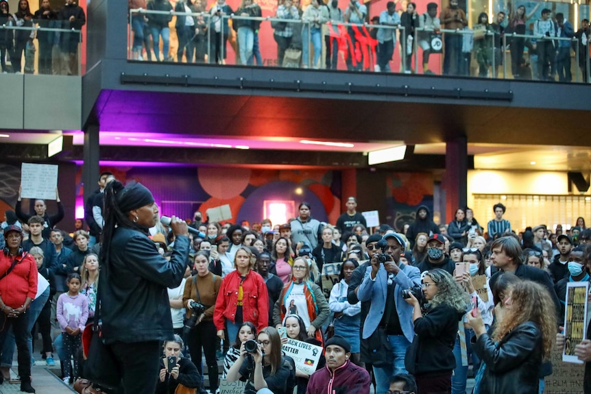 A large crowd, some lining a balcony, listens to an Aboriginal man speaking at a Black Lives Matter protest in the Perth CBD.