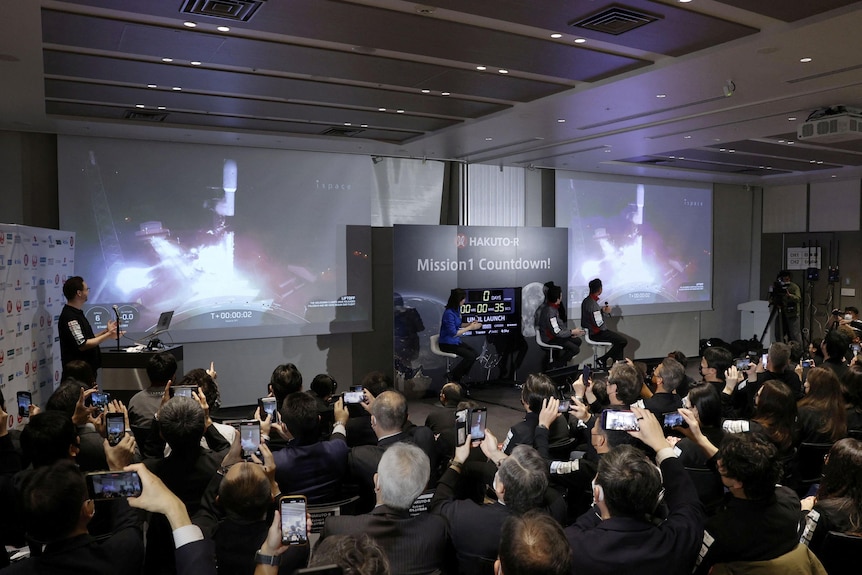 A crowd of people sit in front of a large screen showing the launch while recording it on their phones. 