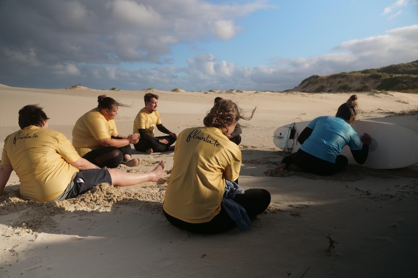 Four young people in wetsuits sitting on the beach in front of a surfboard with therapy messages