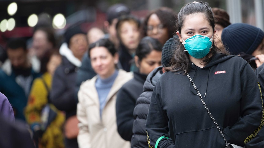 A young woman wears a green face mask and winter clothes standing in a line of people on a street.