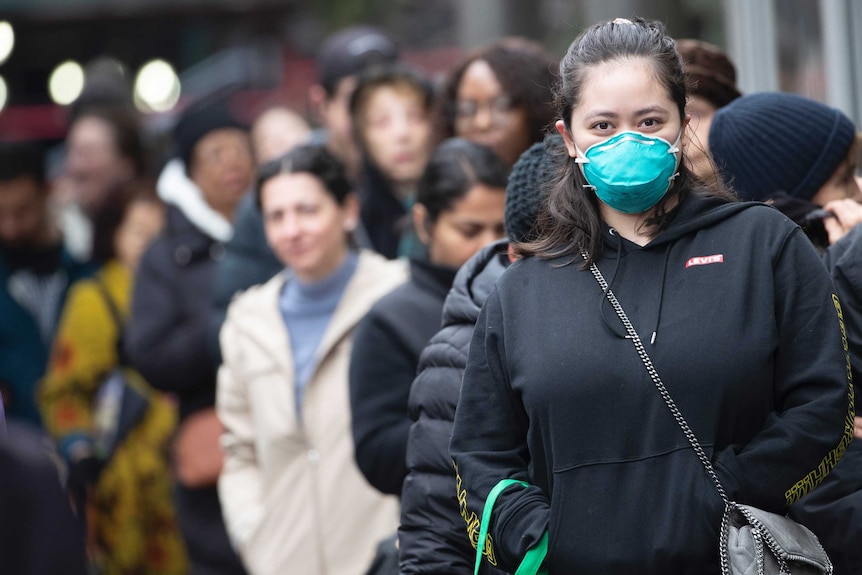 A young woman wears a green face mask and winter clothes standing in a line of people on a street.