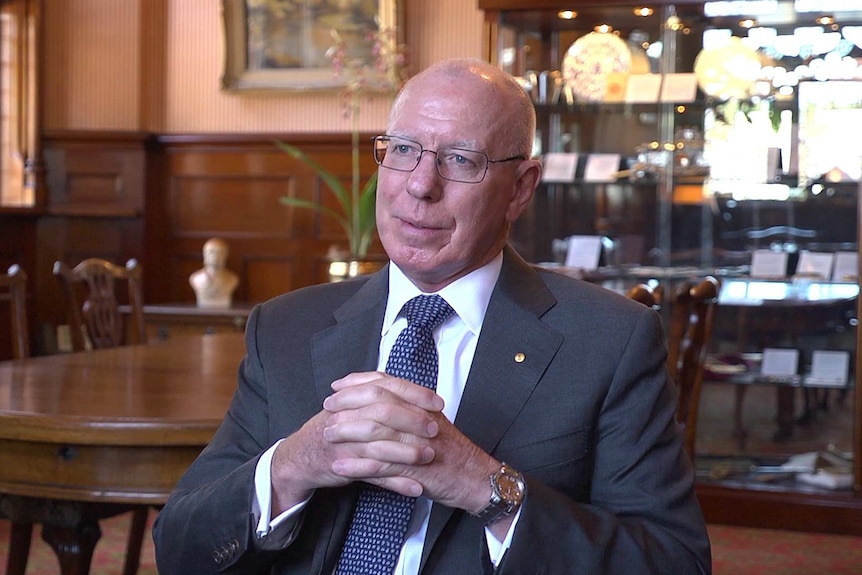 Medium profile shot of a man in a suit sitting in a room and being interviewed off-camera.