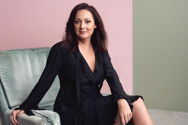 Celeste Barber sits on an armchair in a black dress, smiling at the camera.